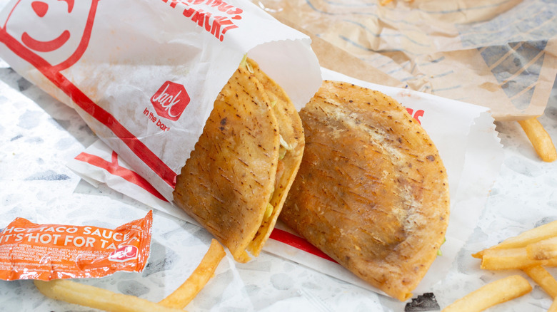 Two Jack in the Box tacos on a paper surface near French fries and hot sauce packets