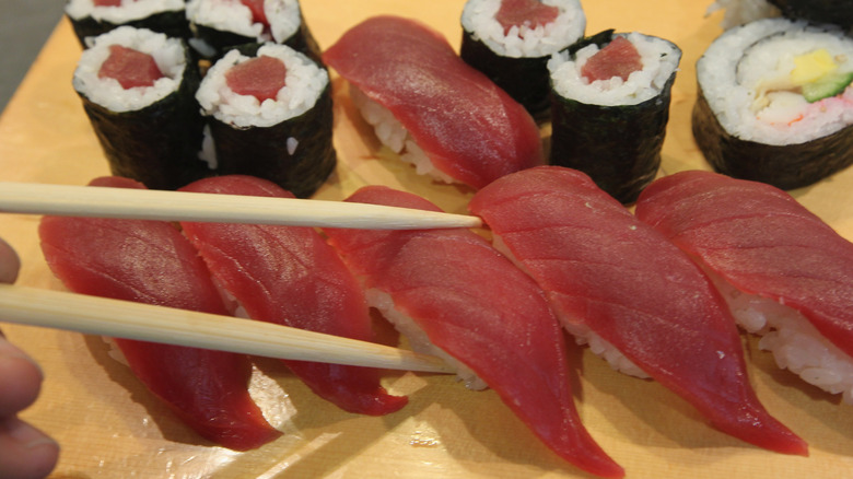 A pair of chopsticks lifts a piece of nigiri off a platter containing various types of sushi