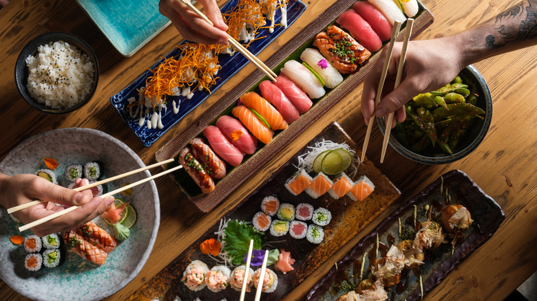 Overhead view of platters of sushi and sashimi with four hands with chopsticks hovering over them