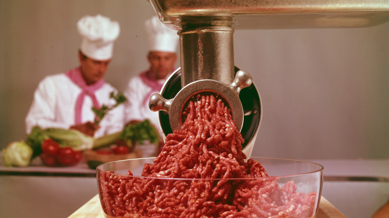 Close-up of uncooked ground beef in a glass bowl under a meat grinder. A pair of male chefs are visible in the background.