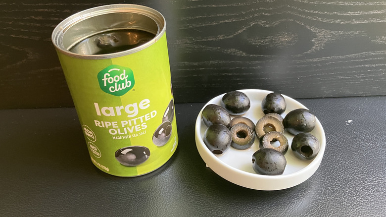 A can of Food Club brand ripe pitted olives and a white dish displaying both whole and sliced black olives.