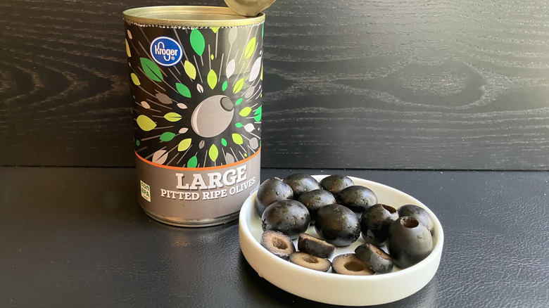 A can of Kroger pitted ripe black olives and a white dish displaying both whole and sliced olives