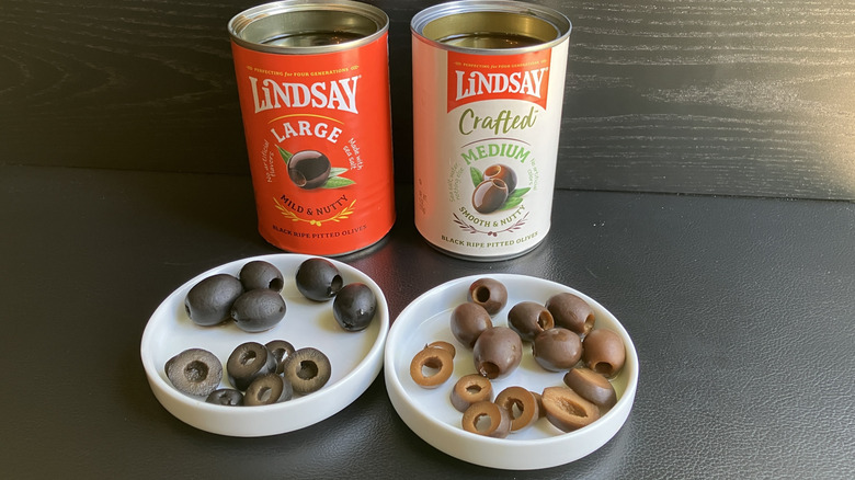 Two white dishes, one displaying Lindsay's black olives and the other showing the olive-green/tan colored Lindsay Crafted olives, with the respective cans in the background