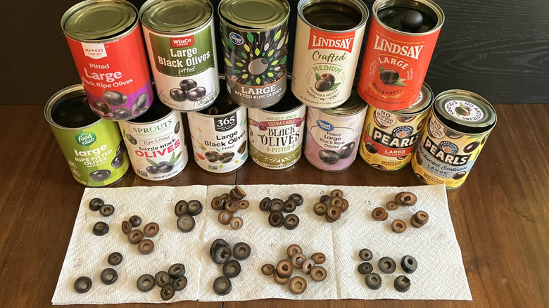 Twelve cans of black olives of varying brands stacked into a wall with piles of sliced olives on a white paper towel in front showing some black and some brown and some green.