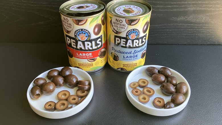 Two cans of Pearls large pitted ripe olives and two bowls displaying both whole and sliced olives which are brownish green in color