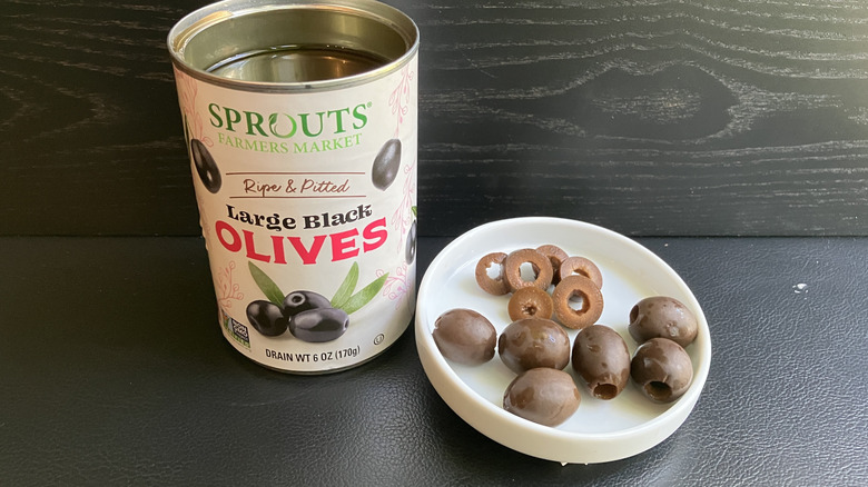 A can of Sprouts brand ripe and pitted large black olives and a white dish displaying both whole and sliced olives. which are a brownish green color.