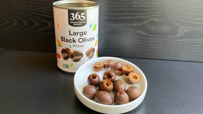 A can of Whole Foods Market 365 brand pitted large black olives and a white dish displaying both whole and sliced olives. which are a brownish red color with tannish-pink insides