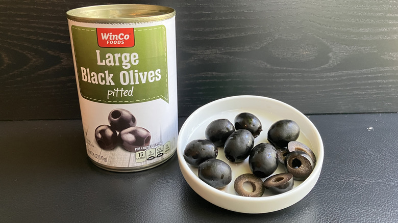 A can of WinCo Foods pitted black olives and a white dish displaying both whole and sliced black olives.
