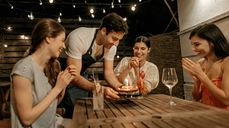 Male waiter in apron delivers birthday cake to table of three women clapping, at outdoor patio restaurant