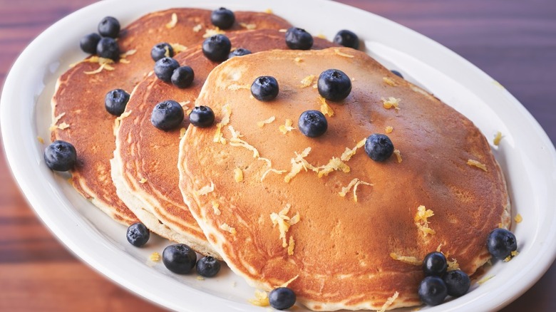 Three pancakes laid out on a a plate, topped with blueberry and lemon zest