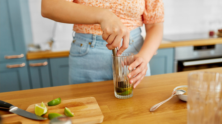 Close-up of woman with pestle and lime in glass making mojito cocktail at home kitchen