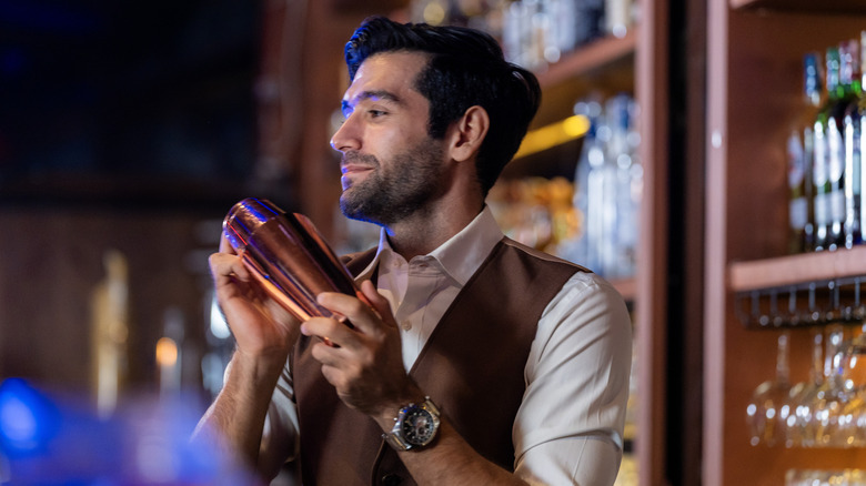 A smiling male bartender in a brown waistcoat mixing a drink in a copper cocktail shaker