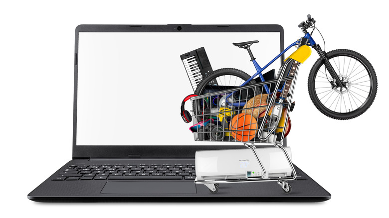 A shopping cart filled with many goods like a bicycle, music instrument, electronics, and tech products set on a laptop.