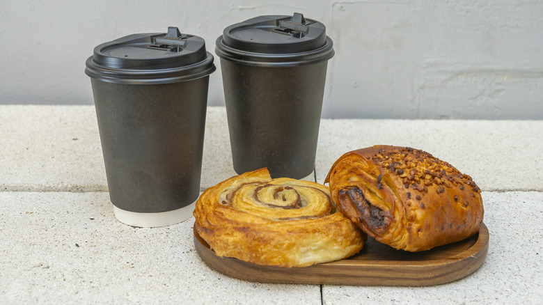 Two black disposable coffee cups with a cinnamon swirl and a chocolate nut pastry on a wooden board over a concrete surface.