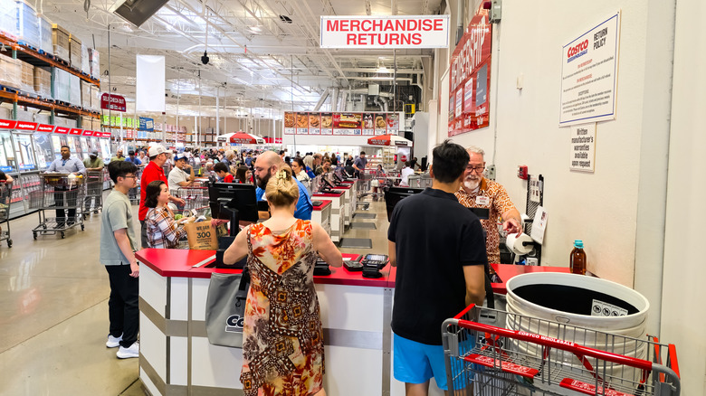 Customers wait in line at the merchandise returns desk inside a Costco warehouse. The store is bustling with activity, showcasing the popularity of Costco's membership-based shopping model.