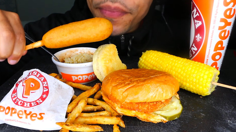 Person holding a Popeyes corn dog alongside a burger, fries, corn, and a drink