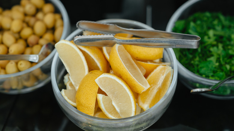 Fresh lemon slices in glass bowl with metal tongs beside salad ingredients on dark buffet counter