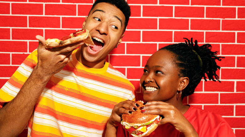 A man eating a big slice of pizza next to a woman with a large burger in front of a red brick wall