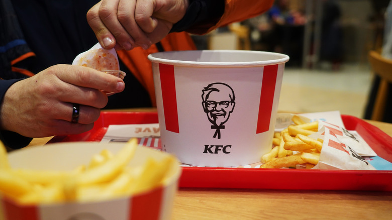 A person's hands opening a container of sauce over a tray with a KFC bucket and a serving of french fries.