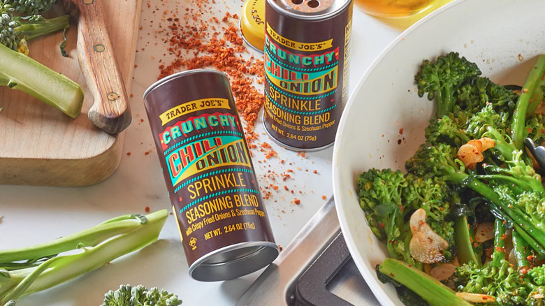 Shaker of Trader Joe's Crunchy Chili Onion Sprinkle Seasoning Blend alongside sauteed broccolini