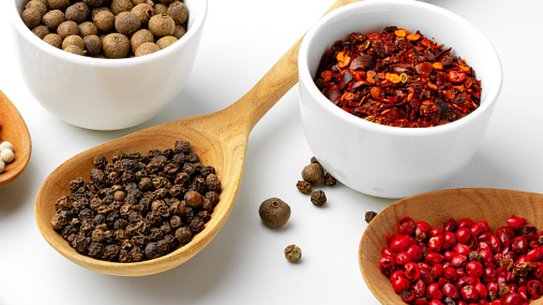 Small white bowls and wooden spoons with black pepper, red pepper flakes, pink peppercorns, and allspice