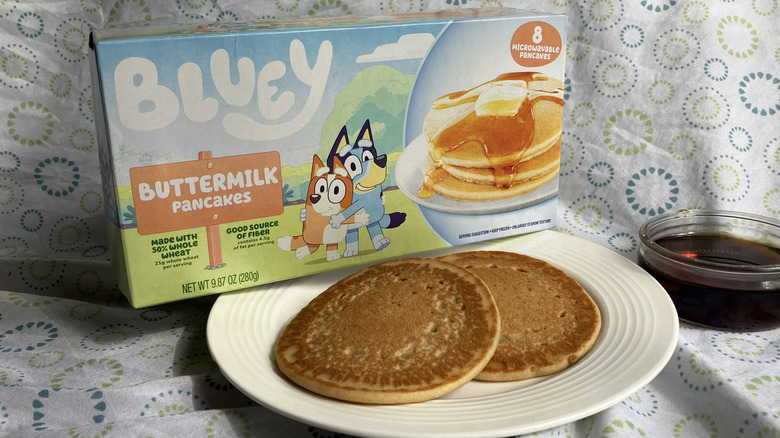 A plate of cooked pancakes next to a glass dish of maple syrup and a box of Bluey brand frozen buttermilk pancakes with Bluey and Bingo on the front.