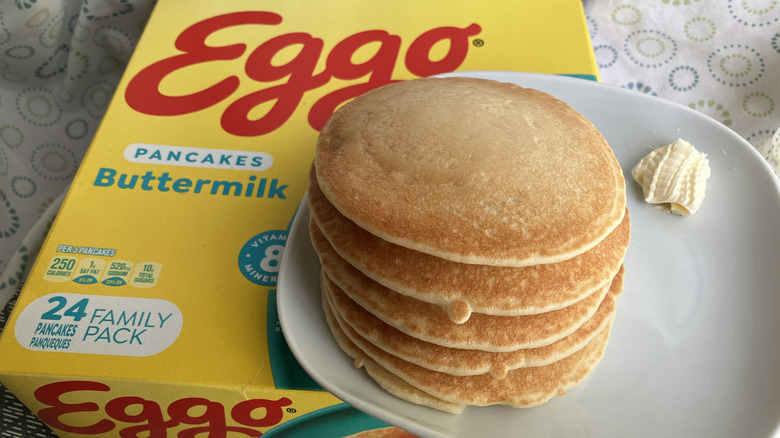 A stack of cooked pancakes on a plate with a dollop of butter, sitting on top of a large yellow box of Eggo brand frozen pancakes