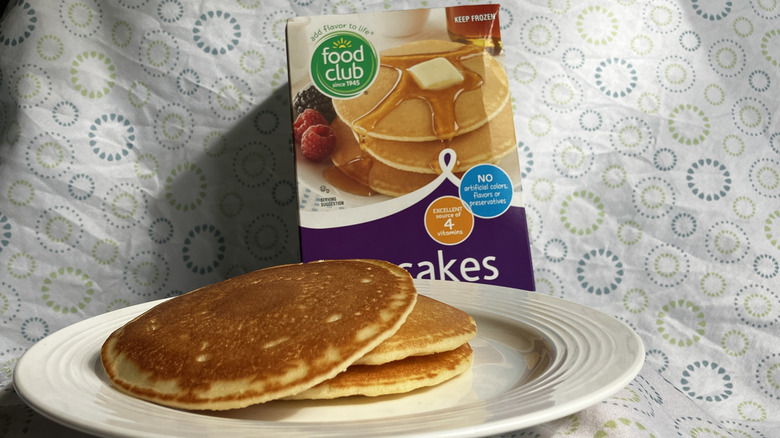 A stack of cooked pancakes on a plate and a box of Food Club brand frozen pancakes