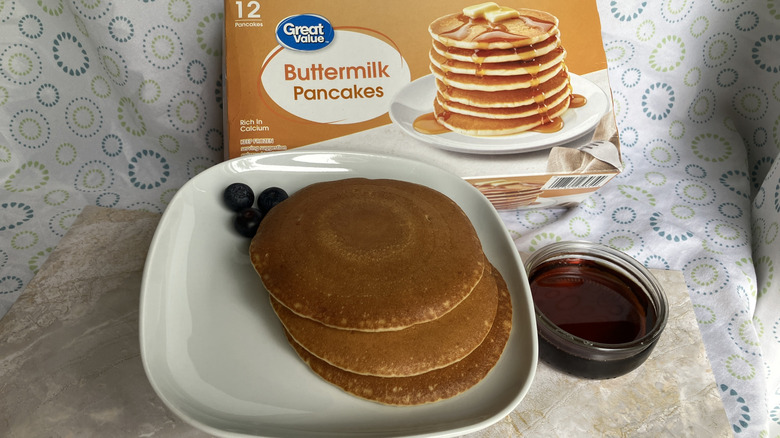 A plate with three cooked pancakes and three blueberries next to a box of frozen Great Value pancakes and dish of maple syrup