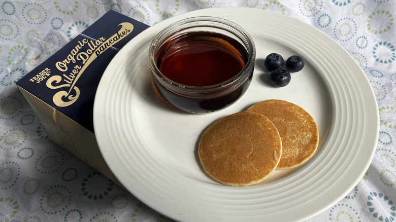 A plate with two mini pancakes, dish of maple syrup, and three blueberries set on top of a box of frozen pancakes