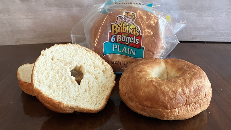 A whole and cut bagel sit in front of a bag of Bubba's Plain Bagels on table.