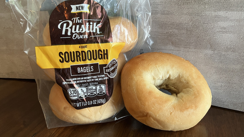 A bagel displayed next to a bag of The Rustik Oven bagels on table.