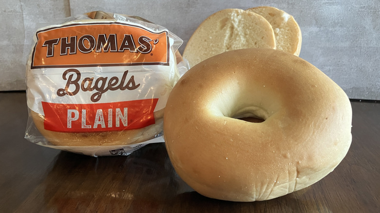 A large bagel sits next to a bag of Thomas' Plain Bagels on table.