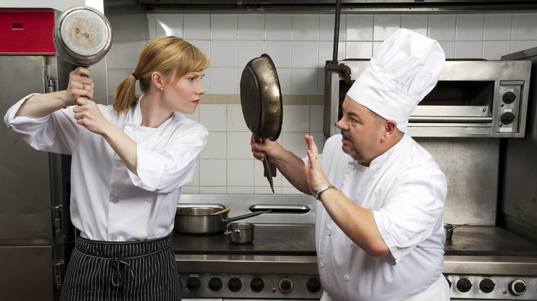 A female and male chef holding pans up at each other in the kitchen
