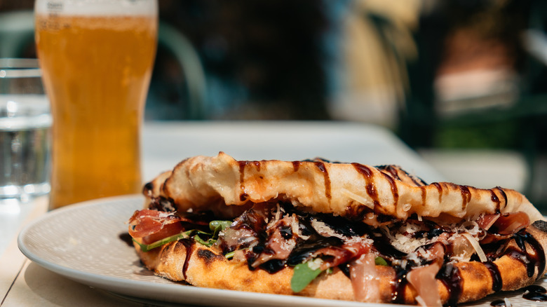 Flatbread sandwich drizzled with balsamic glaze and tall glass of beer in background.