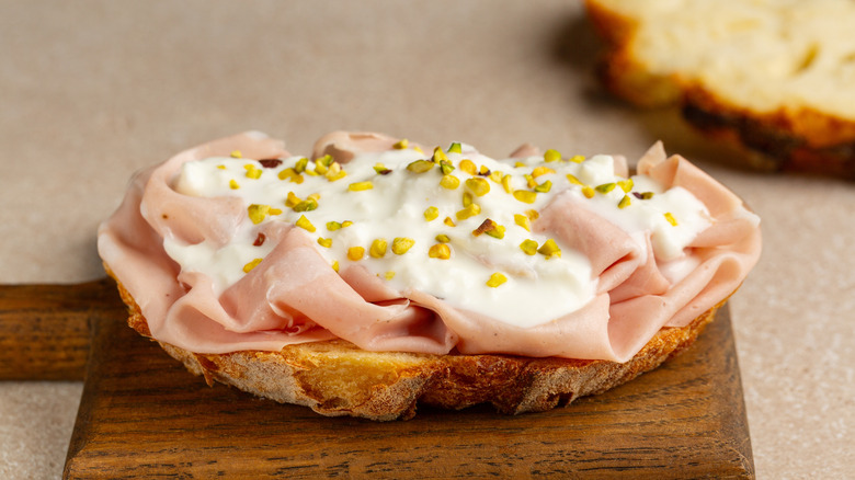 Slice of bread topped with thinly sliced mortadella, creamy cheese, and chopped pistachios.
