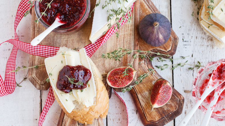 Wooden board holding slice of white bread topped with brie cheese and fig jam alongside fresh figs, fig jam jar, and triangle of brie cheese.