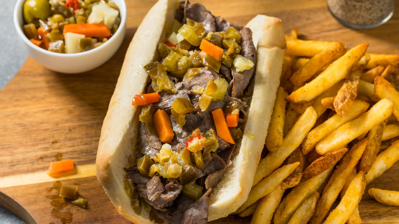 Hoagie bun holding thinly sliced beef topped with giardiniera with fries to the right and a small bowl of extra giardiniera in the upper left.