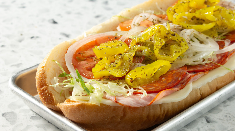 Sub sandwich open with pepperoncini peppers on top of sliced onion, tomato, shredded lettuce, meats, and provolone cheese.