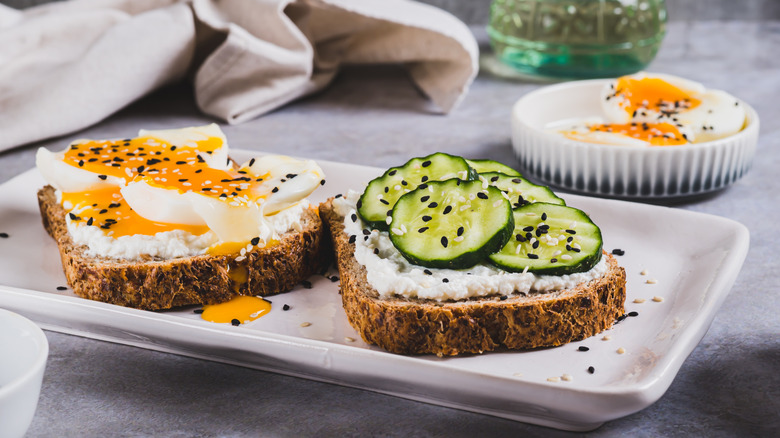 Two slices of toast with ricotta cheese, one topped with soft boiled eggs and one topped with cucumbers.