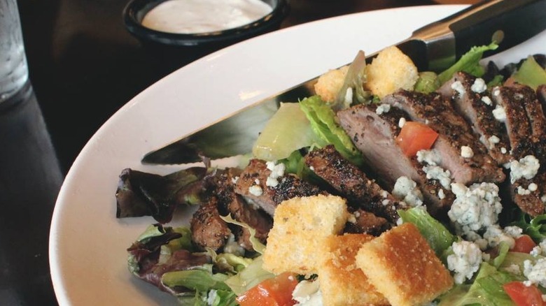 A white platter of salad with a dish of dressing in the background. A knife rests on the plate