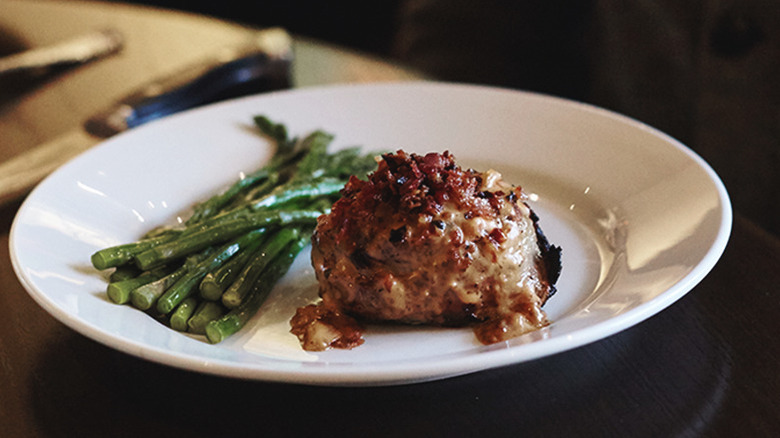 A white plate on a table with a steak covered in sauce and bacon bits with spears of asparagus on the side