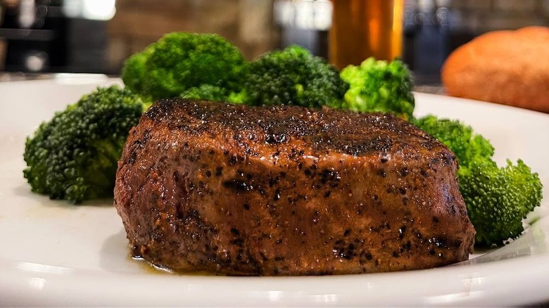 A close up of a filet steak on a white plate with broccoli heads in the background