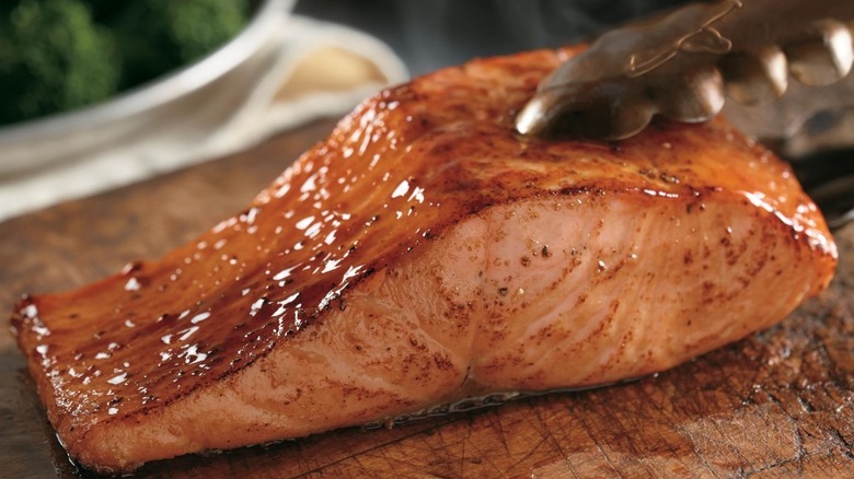 A glistening salmon steak on a wooden platter being held by a pair of tongs