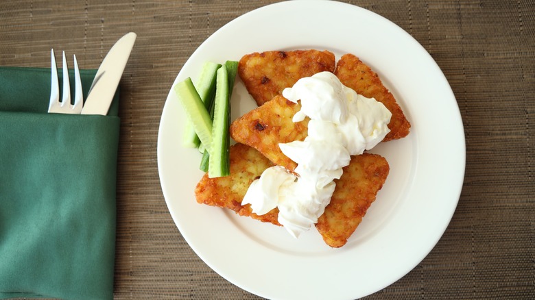 hash browns on a plate with cream on top and sliced cucumbers at the side