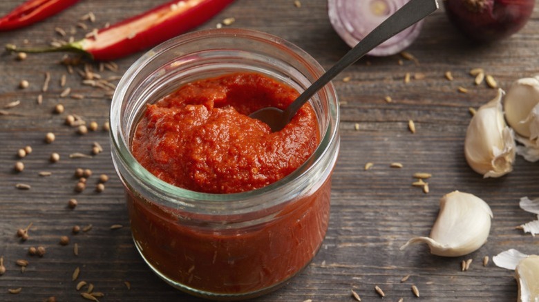 A glass jar of harissa spice paste next to garlic cloves and peppers