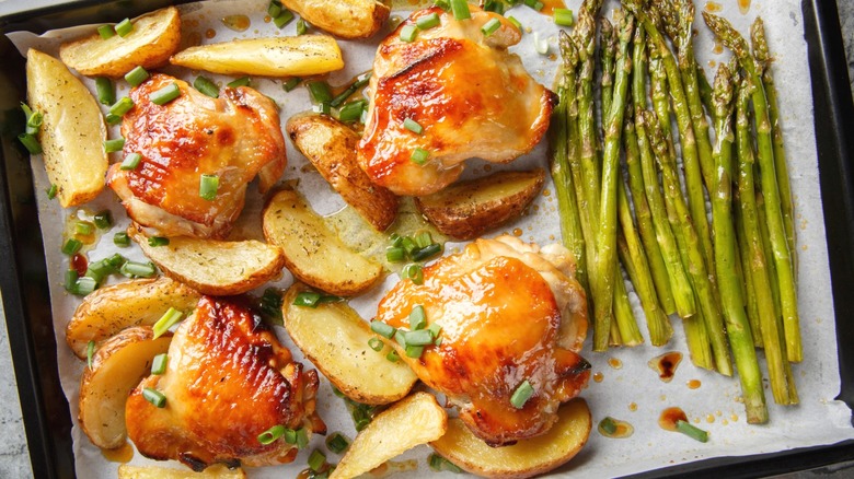 A sheet pan dinner with chicken thighs, potato wedges, and asparagus, with herbs for seasoning.