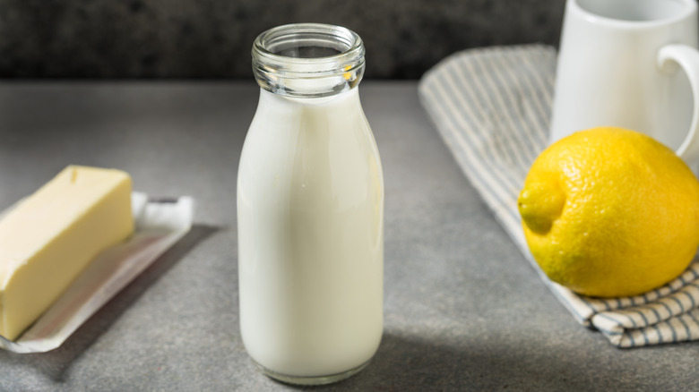 A glass bottle of buttermilk on a countertop flanked by a stick of butter and a lemon