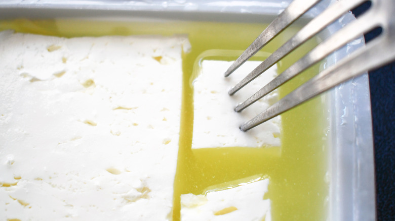 Feta cheese in its brine in a plastic container with a fork sticking into it