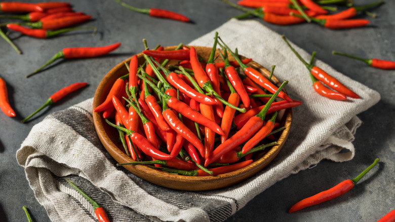Red Thai chili peppers in a wooden bowl over a dish cloth, with more chilis scattered around